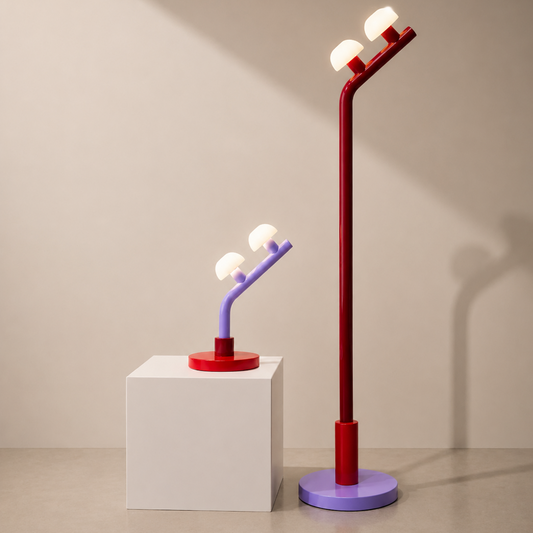 Two modern adjustable floor lamps with dual bulb shades, one in red and purple and one in red and lavender, displayed on a white pedestal and next to it against a warm beige wall.